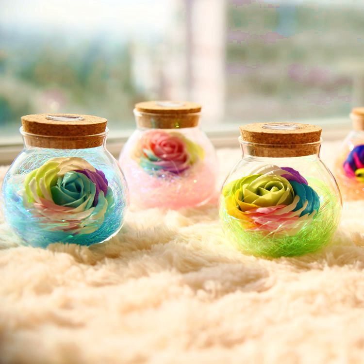 Colorful rose-shaped objects in glass jars with wooden lids on a fluffy surface.