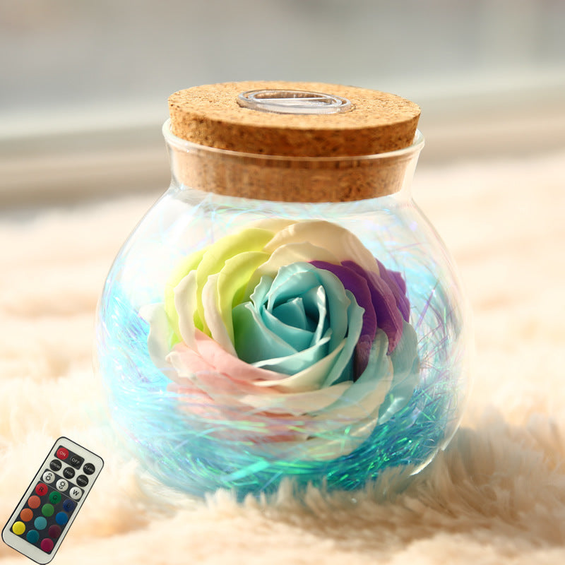 Glass jar with a colorful rose inside, cork lid, on a soft surface with a remote control.