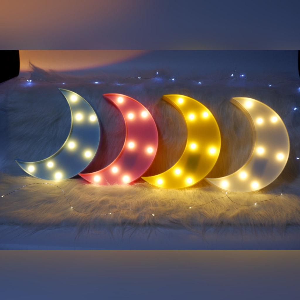 Four crescent moon-shaped lights in blue, pink, yellow, and white on a fluffy surface.