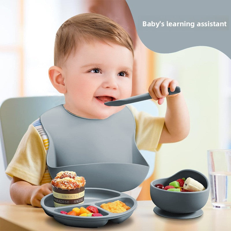 Baby using feeding utensils with a bib and plate, sitting at a table.