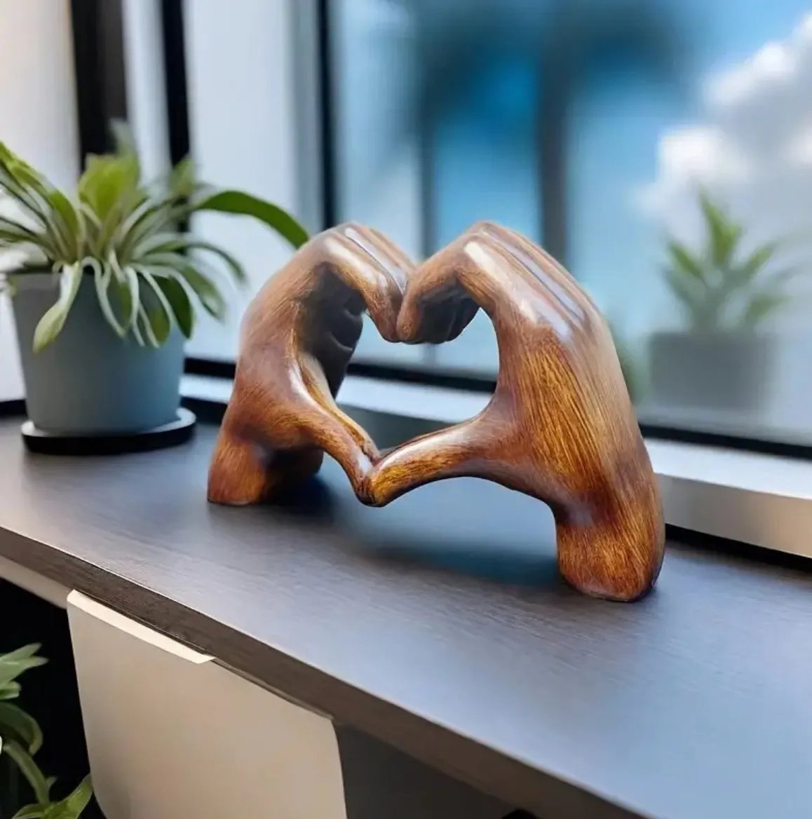 Wooden sculpture of two hands forming a heart shape on a desk with plants in the background