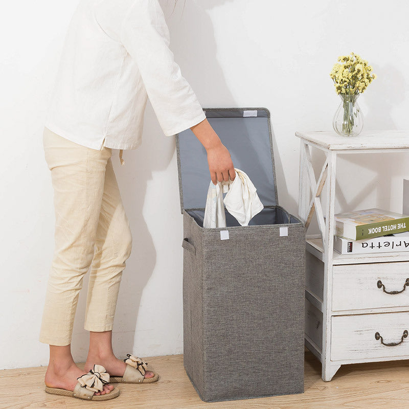 Person holding a white garment over a gray laundry basket in a room with a white wall and wooden floor.