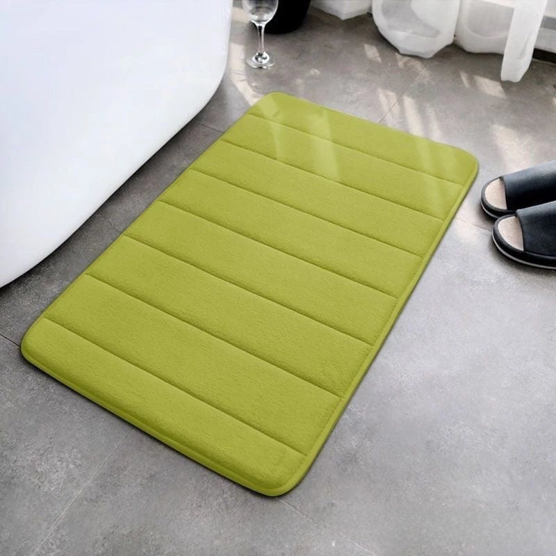 Green bath mat on a bathroom floor with a white bathtub and slippers in the background.
