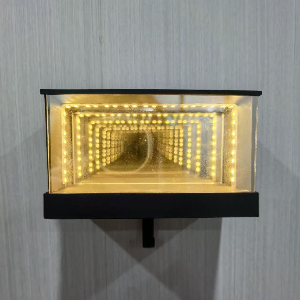 Wall-mounted light fixture with glass cover and black frame on a gray wall.