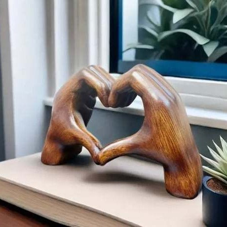 Wooden sculpture of intertwined hands on a surface with a window and plant in the background