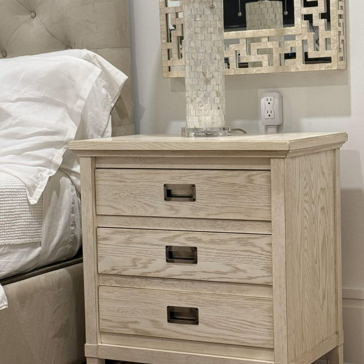 Wooden nightstand with three drawers next to a bed in a bedroom setting.