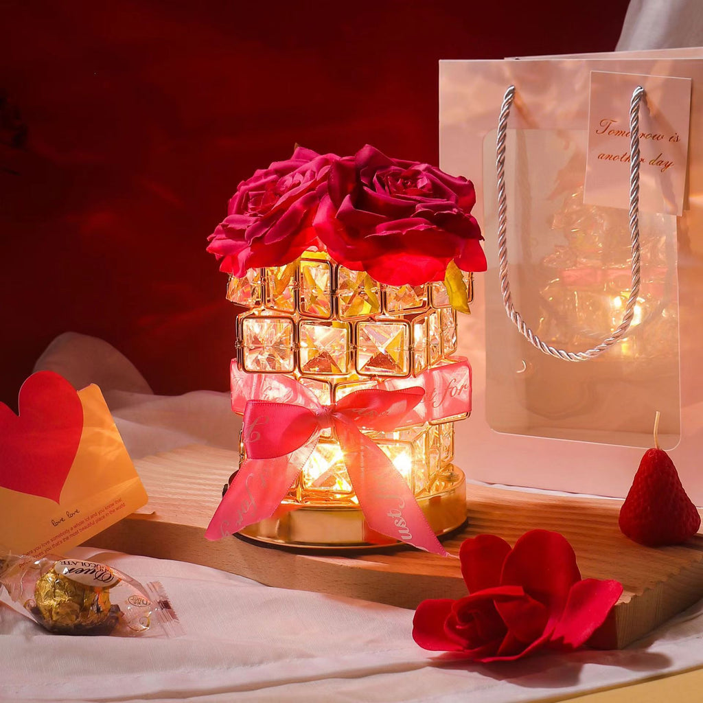 Decorative lamp with red roses, candy, and a gift bag on a wooden surface.