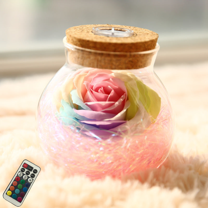 Glass jar with cork lid containing a colorful rose and LED lights, with a remote control on a soft surface.