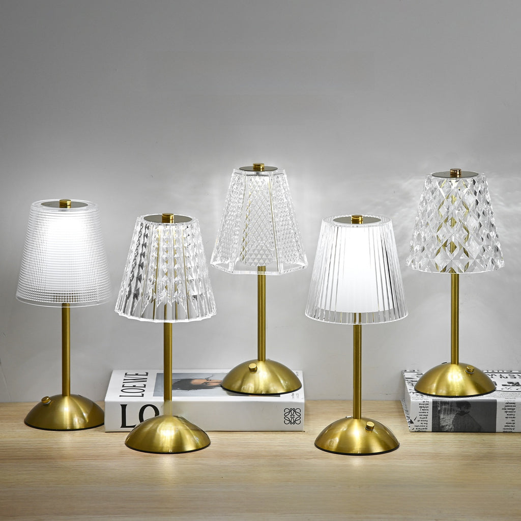 Five table lamps with gold bases and white, clear, and patterned shades on a wooden surface.