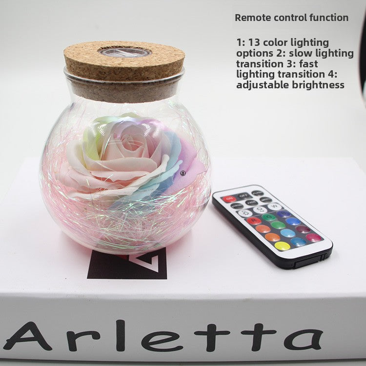 Decorative LED rose in a glass jar with cork lid and remote control on a white surface.